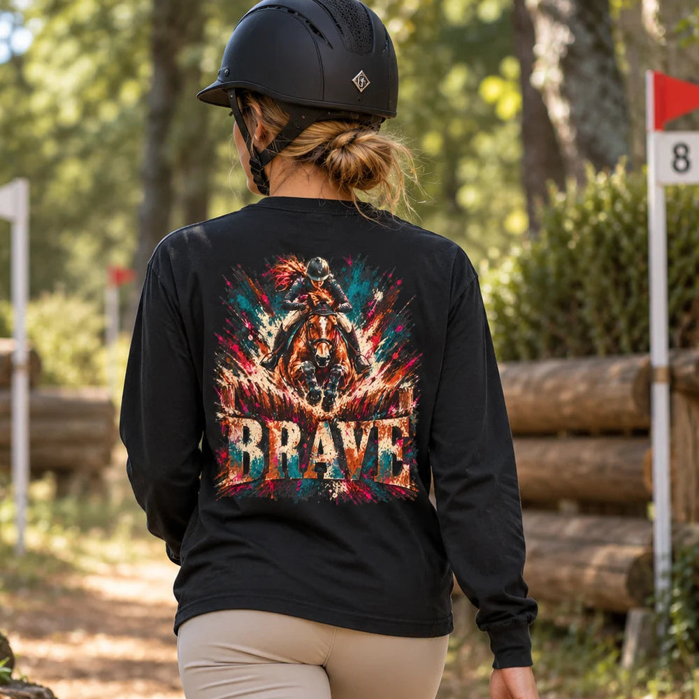 A woman wearing a black long-sleeve equestrian shirt with a large colorful 'BRAVE' graphic on the back, riding boots, and a black riding helmet, shown from behind on an outdoor horse show track.